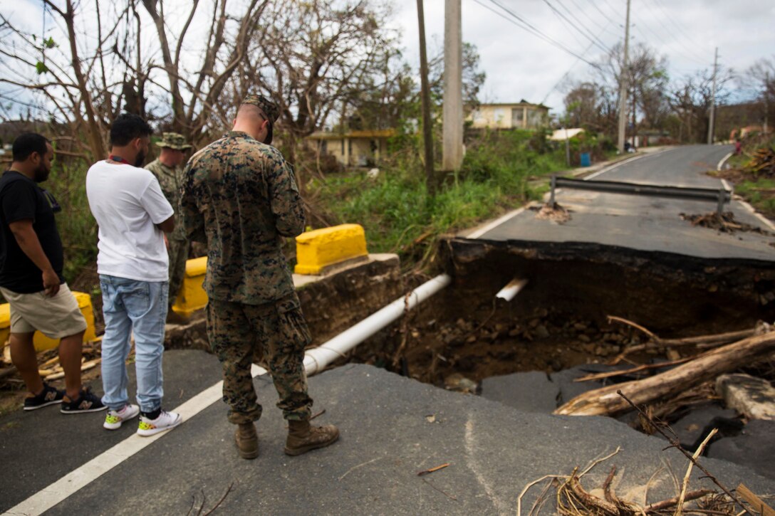 A marine and civilians stand next to a damaged road.