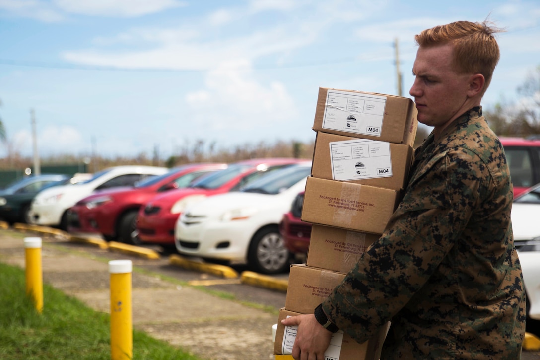 A sailor carries boxes through a parking lot.