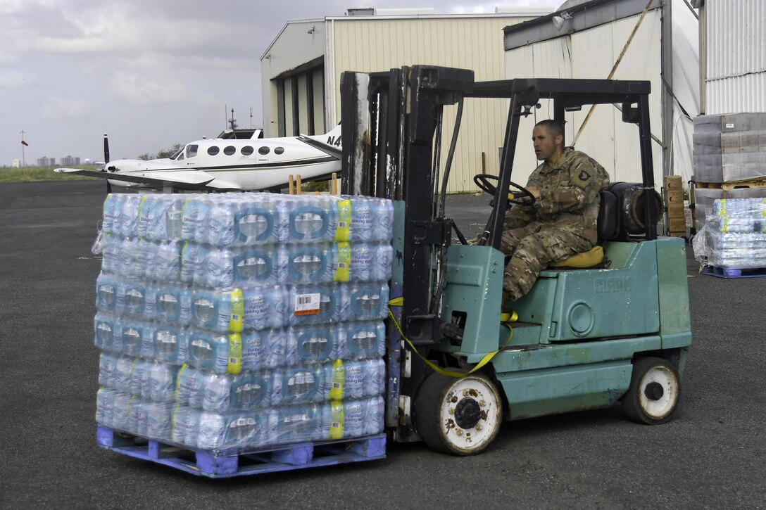 A soldier sits at the controls of a forklift moving cases of bottled water.