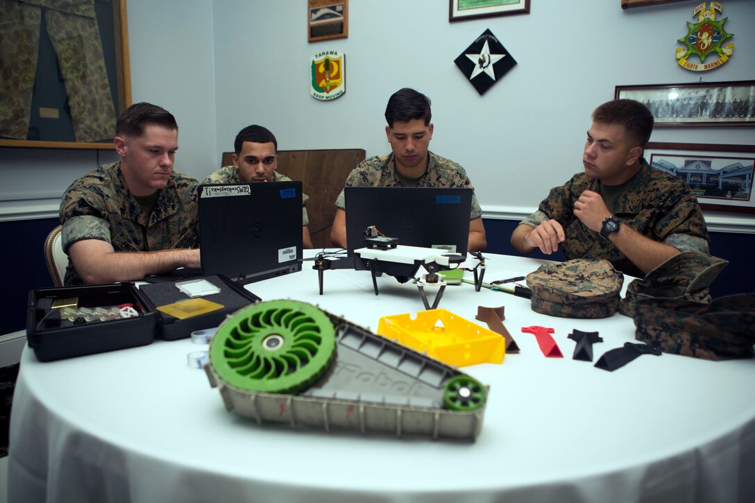 Four Marines sit at a table looking at computers with objects on the table.