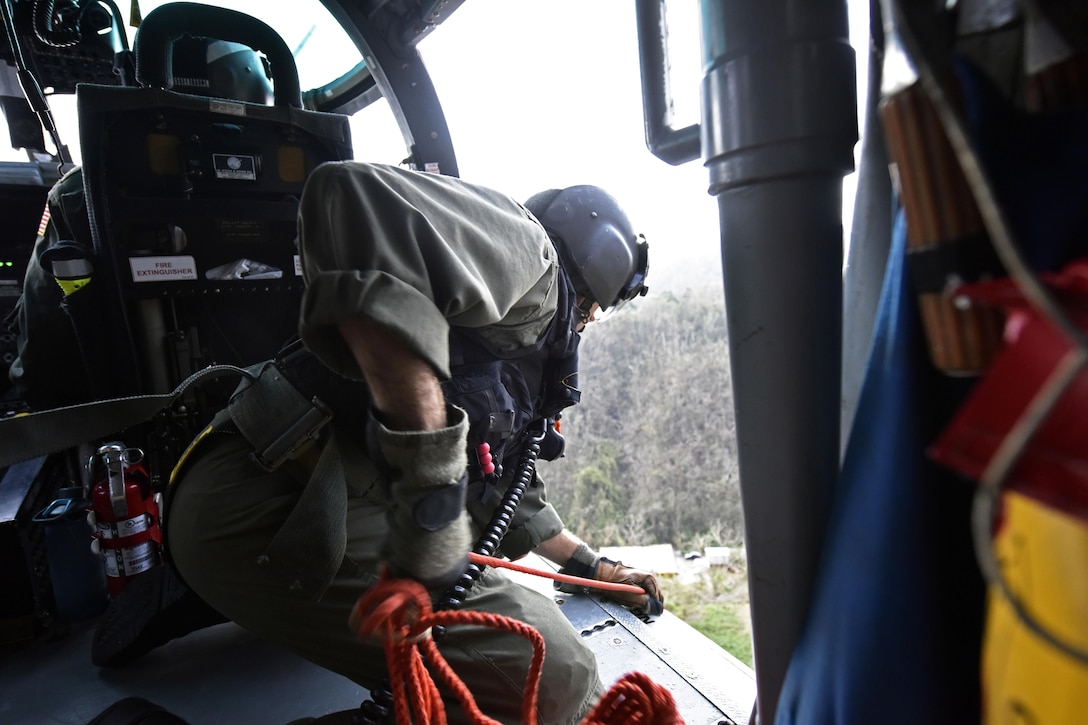 A Coast Guardsman leans toward a helicopter door holding a rope.