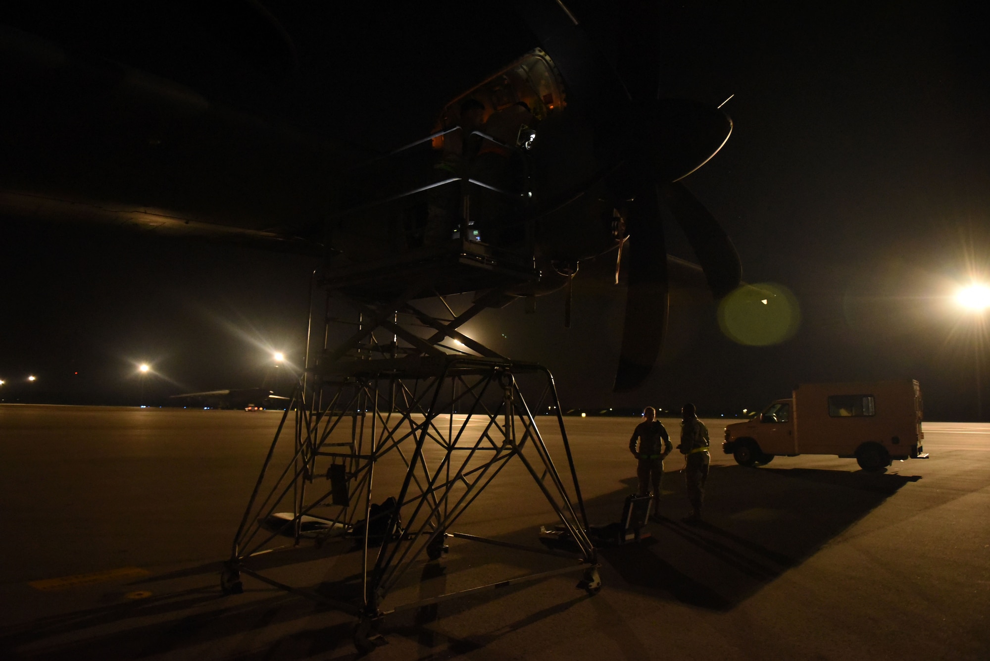 Two people on a maintenance stand work on an airplane prop on a flight line at night.
