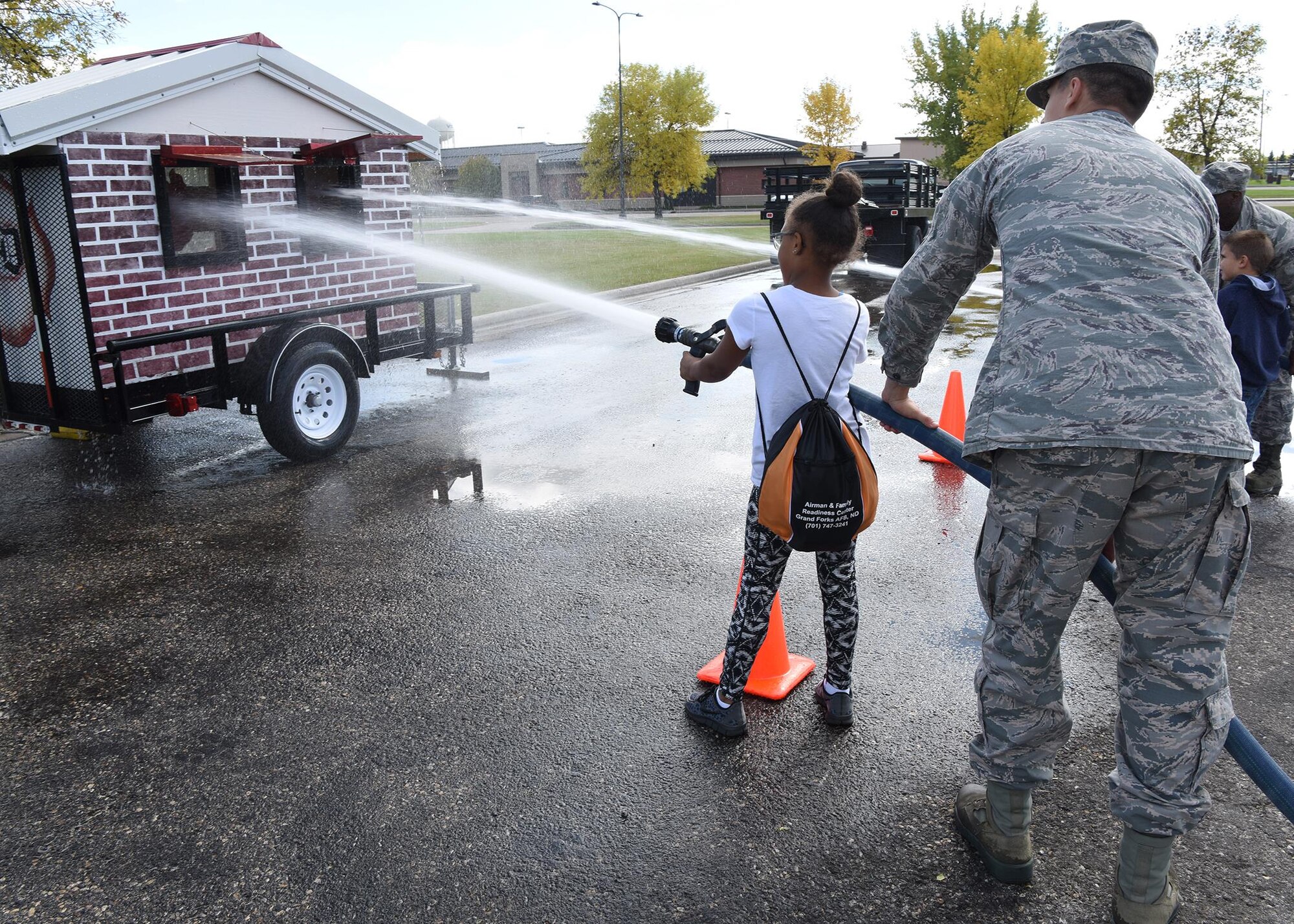 Staff Sgt. Jordan Patterson, 319 Civil Engineer Squadron fire inspector, helps a child spray water into a play house during a kid’s deployment day event Sept. 29, 2017, at Grand Forks Air Force Base, North Dakota. The deployment day, which was sponsored by the Network 5/6, featured activities such as a military working dog demo, obstacle course, combatives and more. (U.S. Air Force photo/Senior Airman Cierra Presentado)