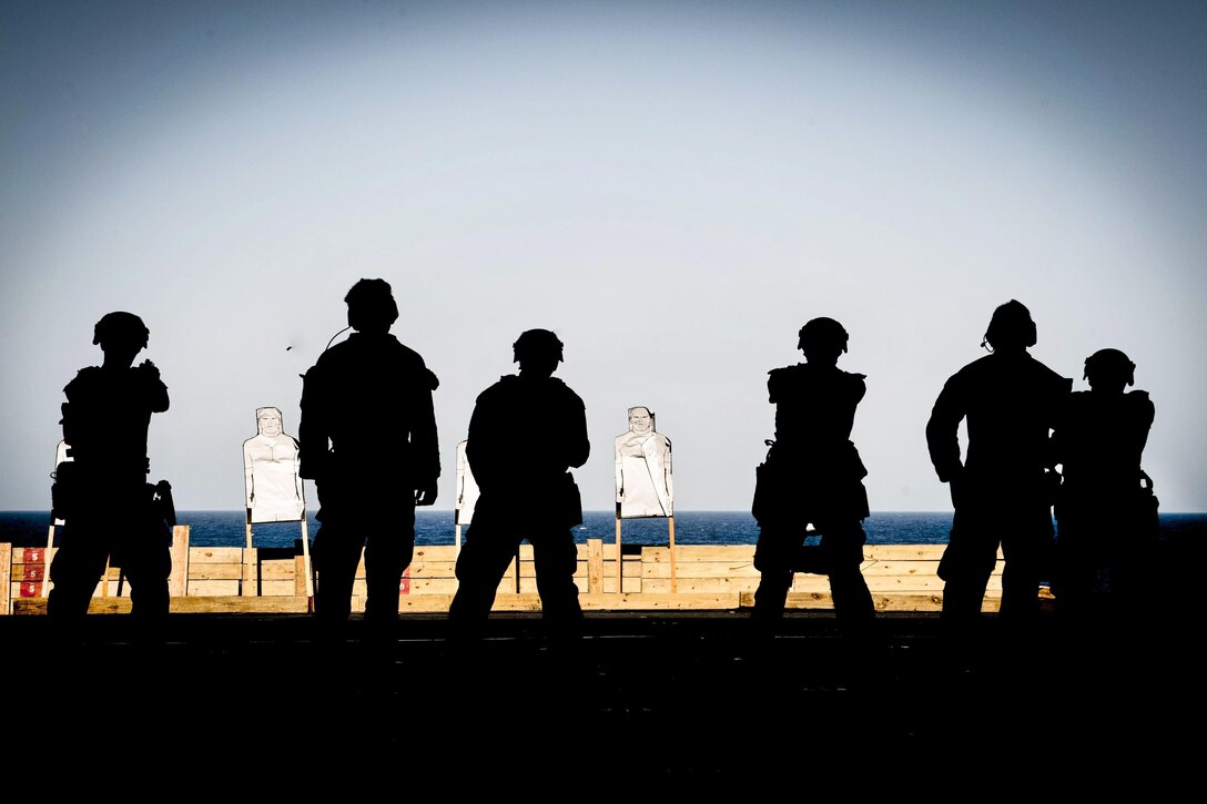 Marines, shown in silhouette, fire at targets on a ship's deck.