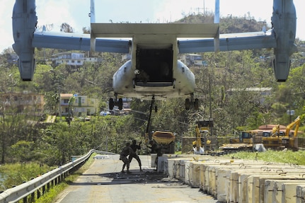 Marines standing on a roadway sling-load a barrier to a low-flying aircraft.
