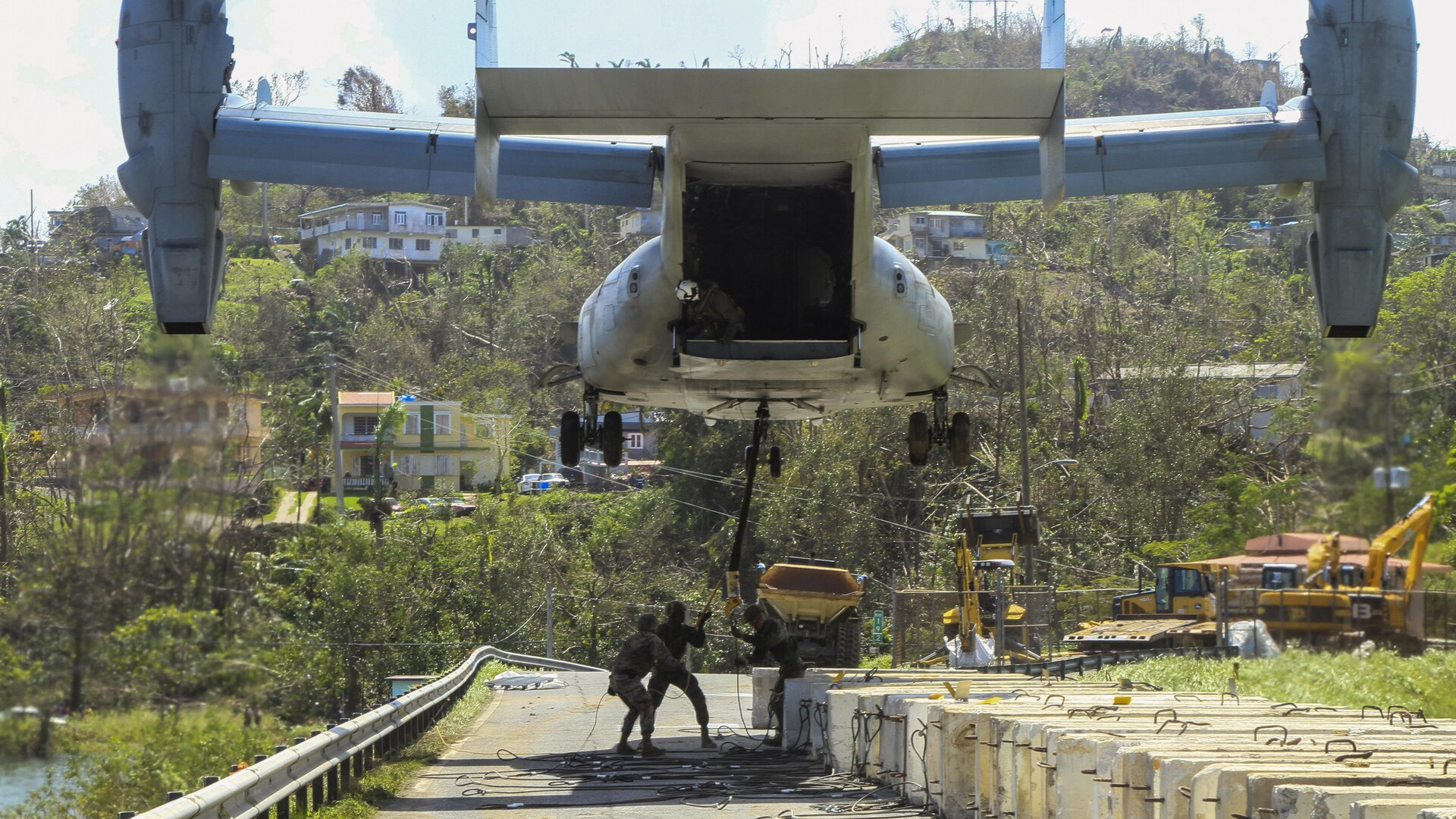 Marines standing on a roadway sling-load a barrier to a low-flying aircraft.