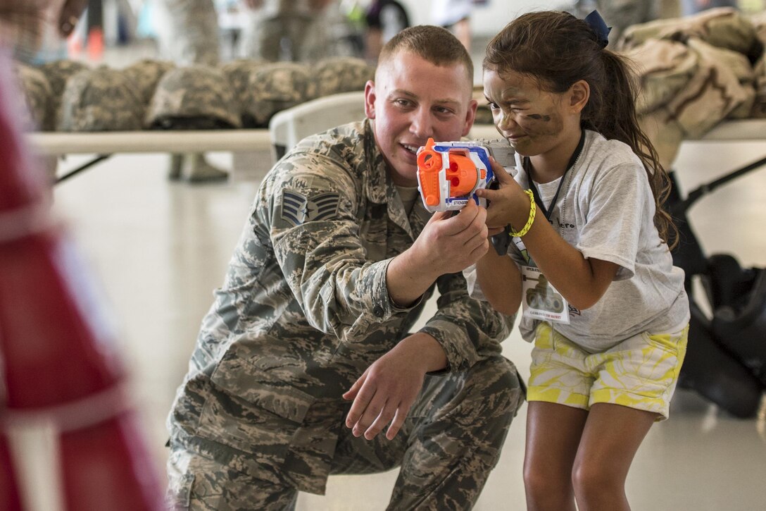 A girl holds a foam gun and squints as an airman kneels and helps her.