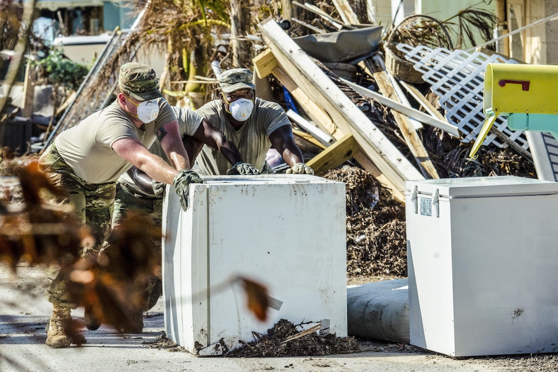 Two soldiers wearing dust masks push a refrigerator outside amid debris.