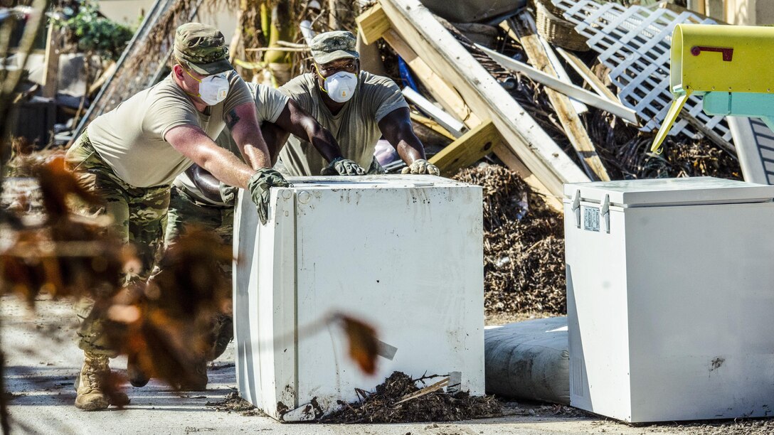 Two soldiers wearing dust masks push a refrigerator outside amid debris.