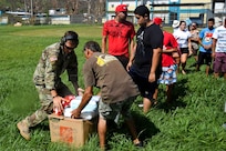 A soldier and civilian unload boxes while a line of people waits.