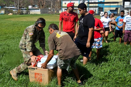 A soldier and civilian unload boxes while a line of people waits.