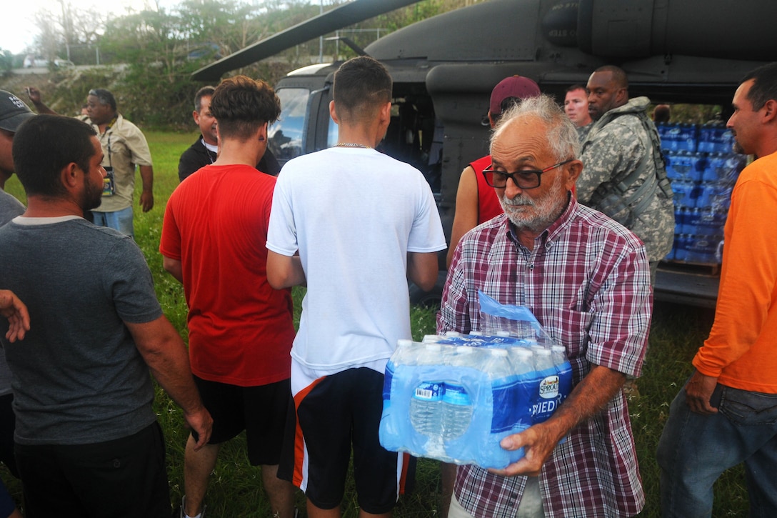 A guardsman hands cases of water to civilians from a helicopter.