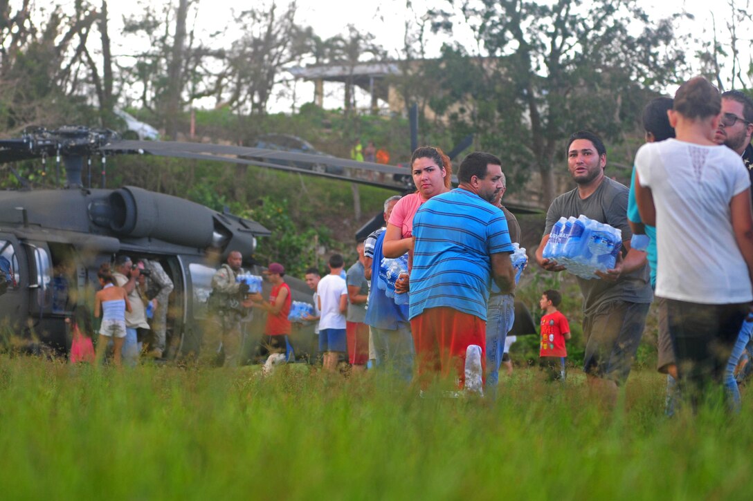 A line of civilians unload cases of water from a helicopter.