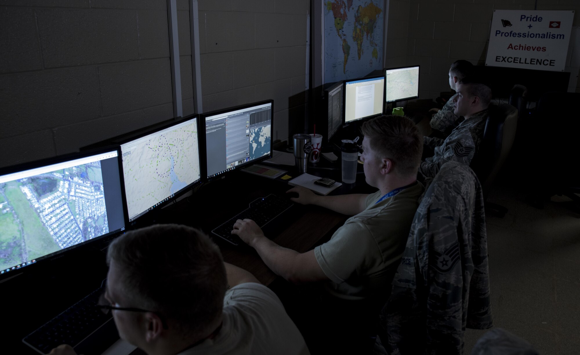 Four Airmen sit in a dark room looking at computer monitors that have over head imagery of locations in Puerto Rico. There is a sign in the background that reads "Pride + Professionalism achieves excellence".