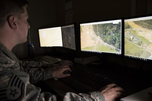 In a dark room, Staff Sgt. Danny sits at a desk looking at two computer screens that have over head images of the Guajataca dam.  You can see the erosion of the dam in the images.