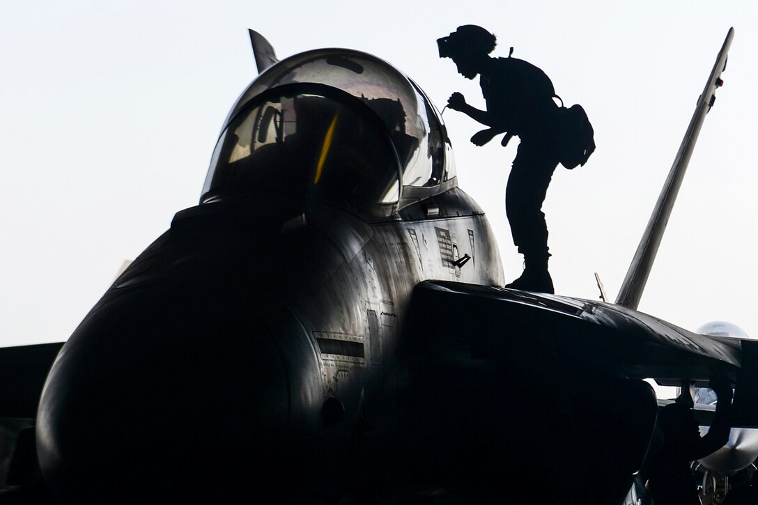 A sailor, shown in silhouette, stands on an aircraft wing and performs maintenance.