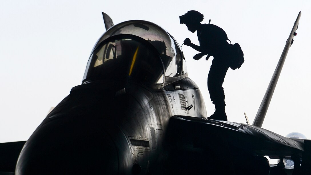 A sailor, shown in silhouette, stands on an aircraft wing and performs maintenance.