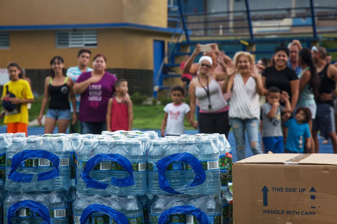 Two groups of people cheer as cases of food and supplies are stacked up.