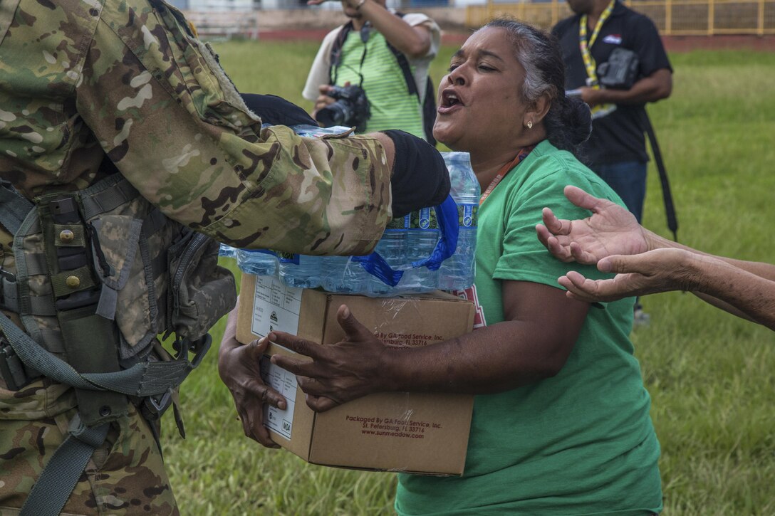 A soldier hands water and a box of relief supplies to a woman.