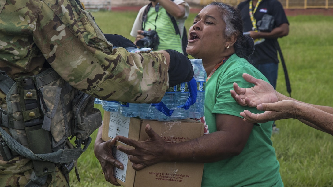 A soldier hands water and a box of relief supplies to a woman.