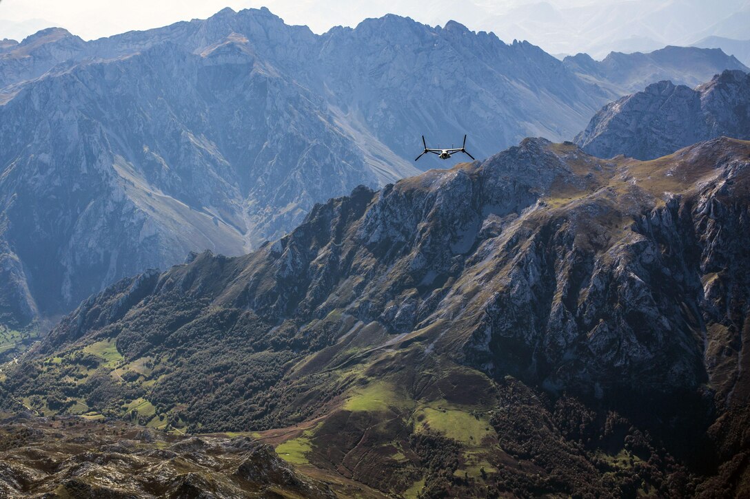 A tiltrotor aircraft flies amid rugged mountain peaks.