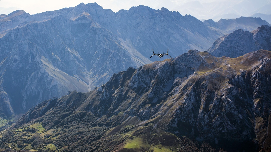 A tiltrotor aircraft flies amid rugged mountain peaks.