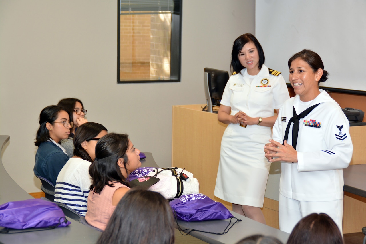 Two sailors talk with students at desks in a classroom.