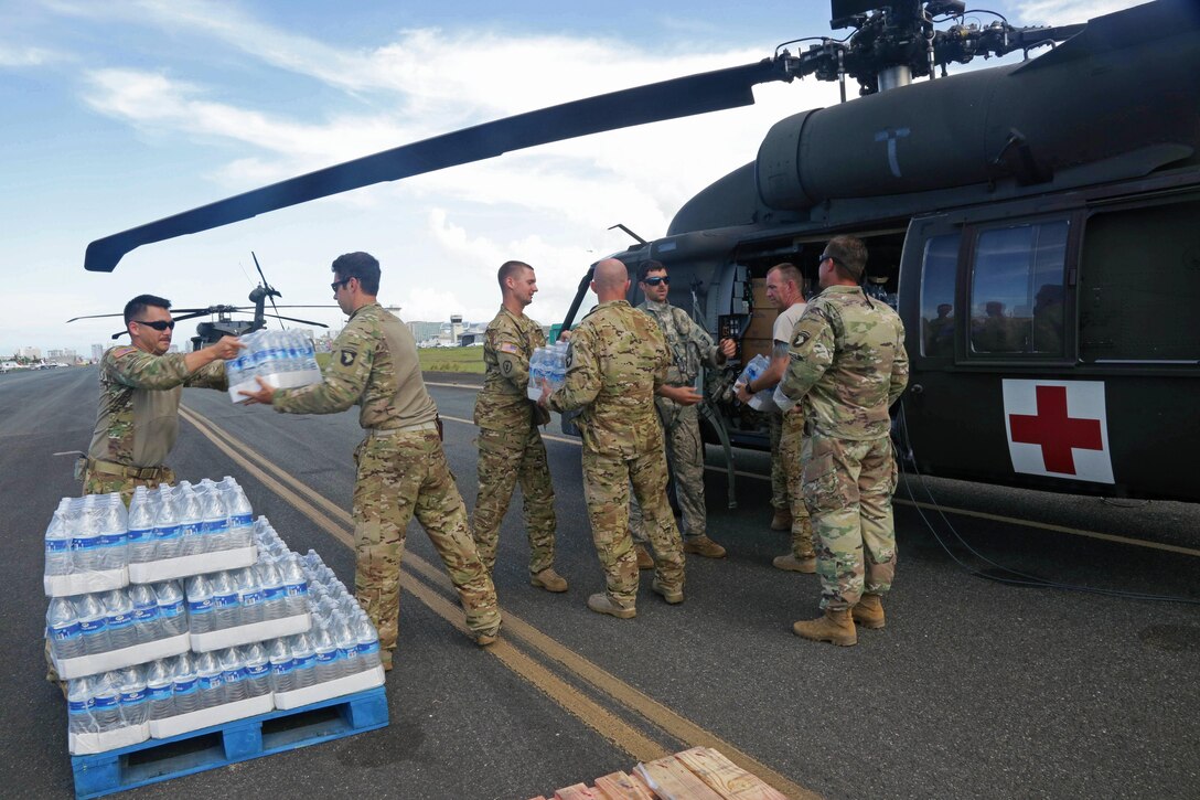 A group of service members hand off water cases to each other next to a helicopter.