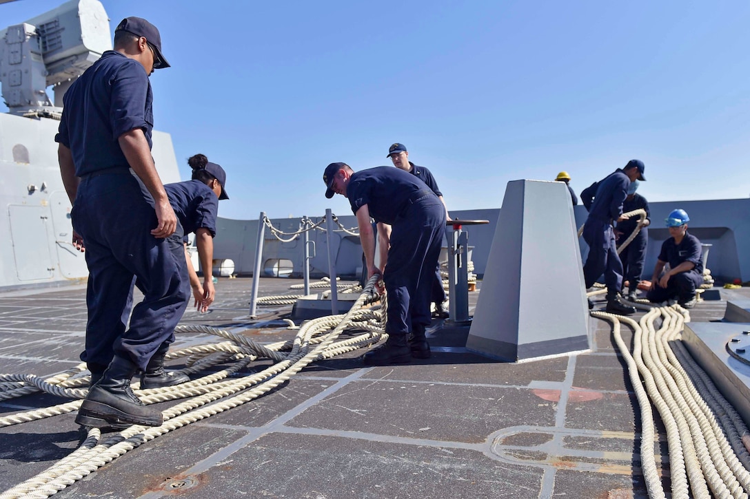 A group of sailors inspect mooring lines on the deck of a ship.