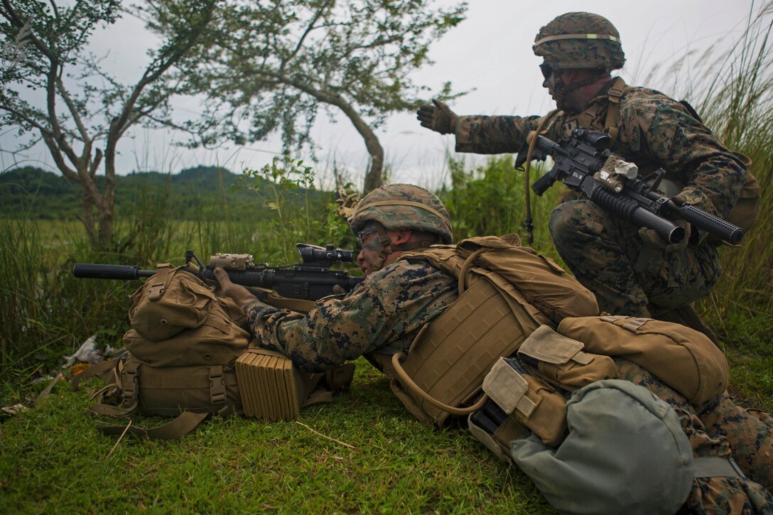 A Marine gestures ahead for another Marine aiming a weapon.
