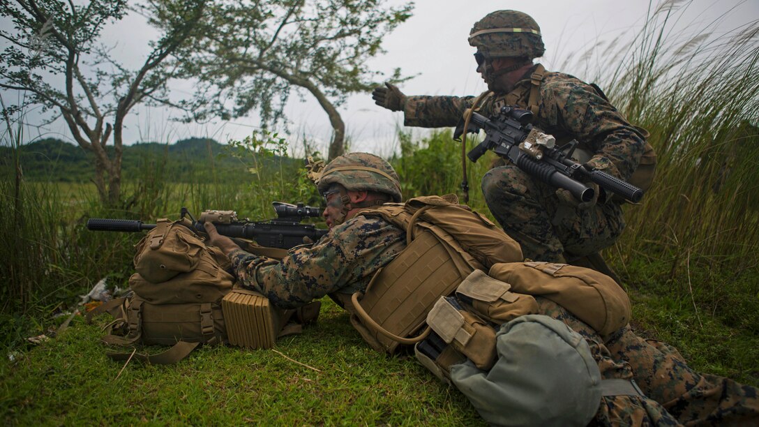 A Marine gestures ahead for another Marine aiming a weapon.