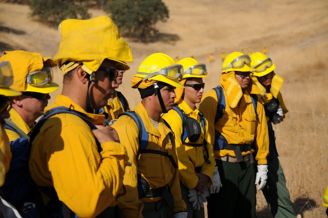 Firefighters wearing yellow shirts and helmets stand together outside while listening to a briefing.