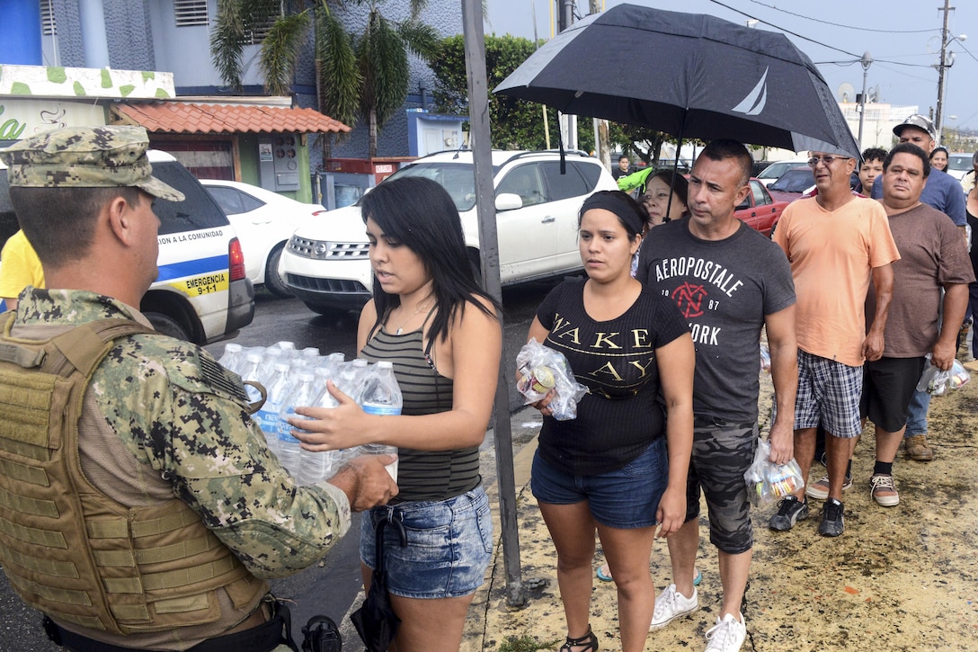 A service member delivers a case of bottled water to a woman standing in line.