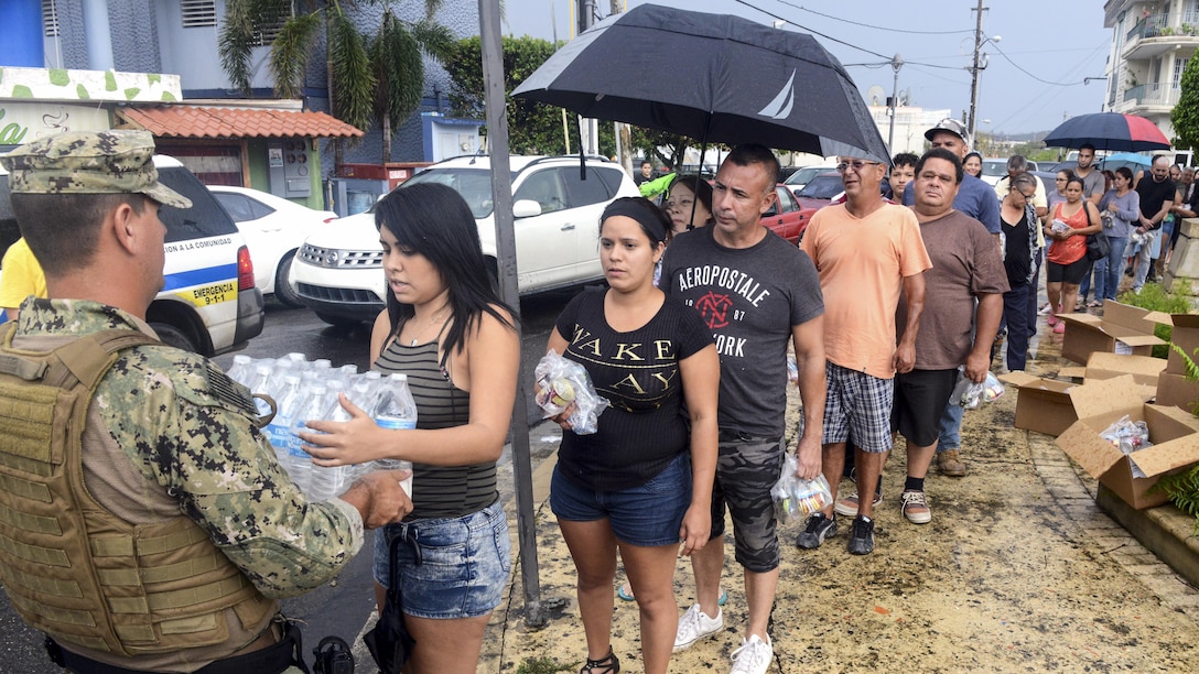 A service member delivers a case of bottled water to a woman standing in line.