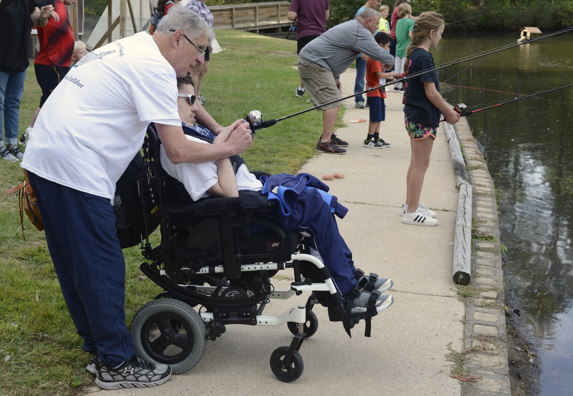 A man helps his wheel-chair bound son fish by a lake, with other people fishing in the background.