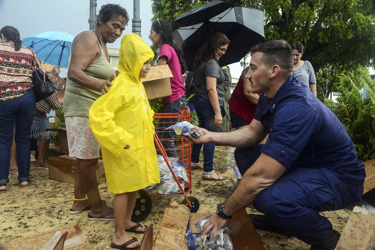 A service member hands a child in a yellow coat some food.