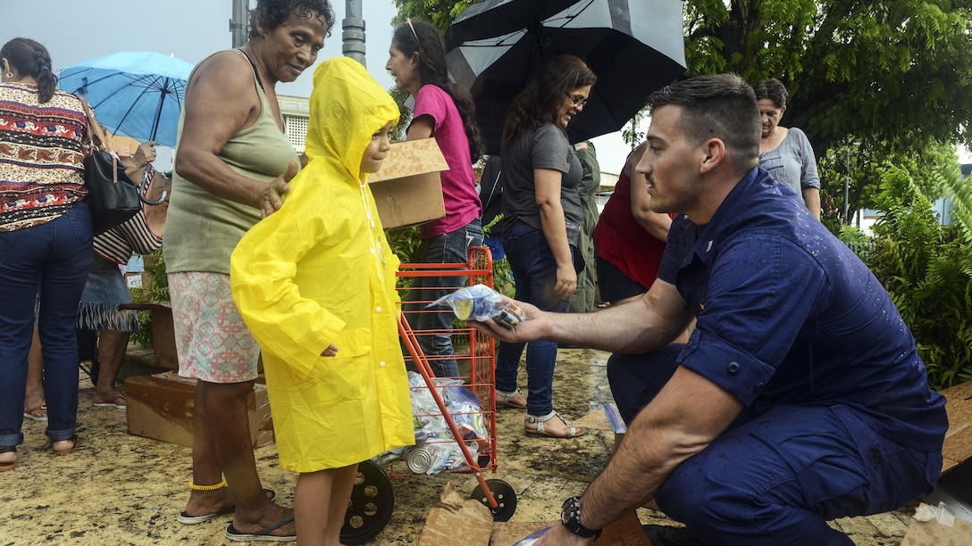 A service member hands a child in a yellow coat some food.