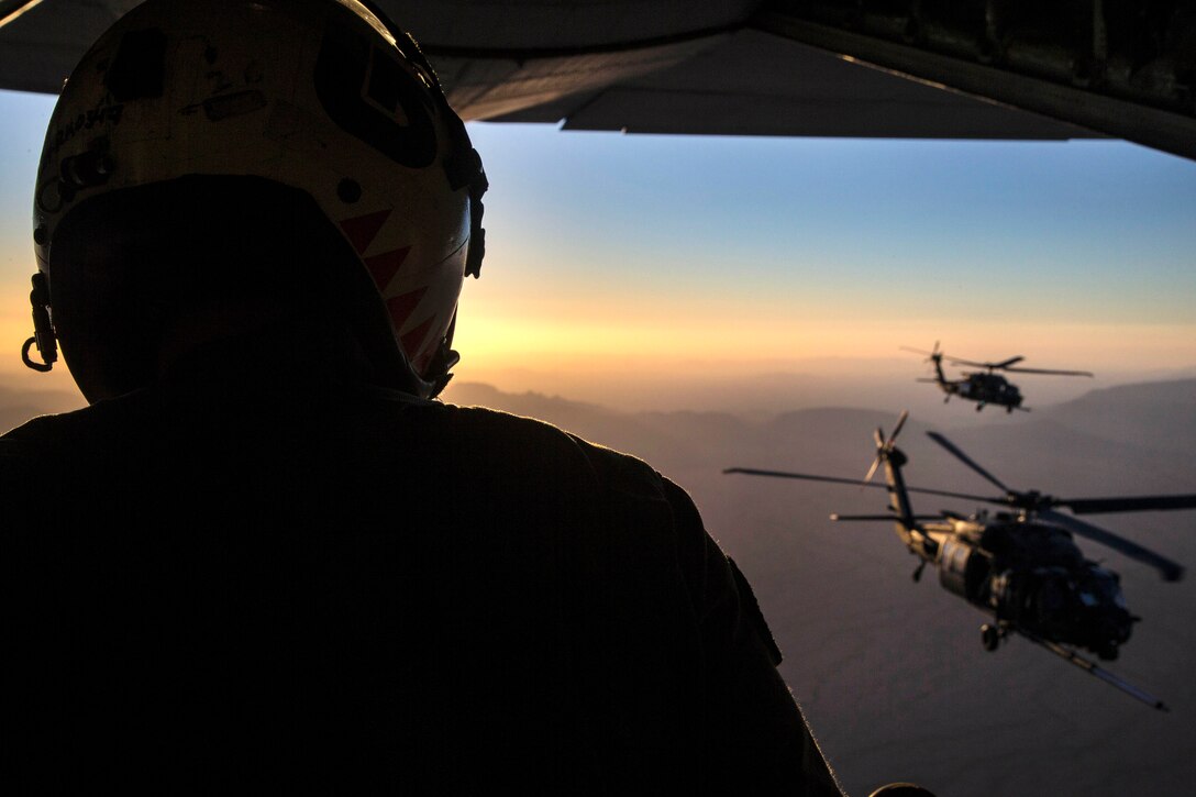 A Marine looks out the open door of a helicopter toward two other helicopters in flight.