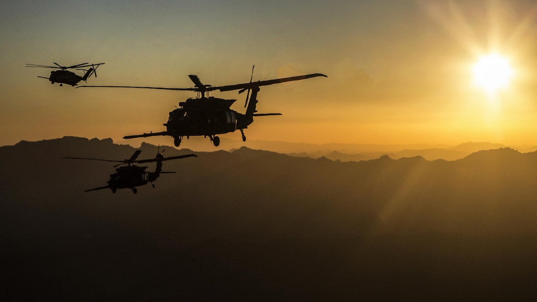 Three helicopters prepare for a refueling exercise with a low sun over the mountains.