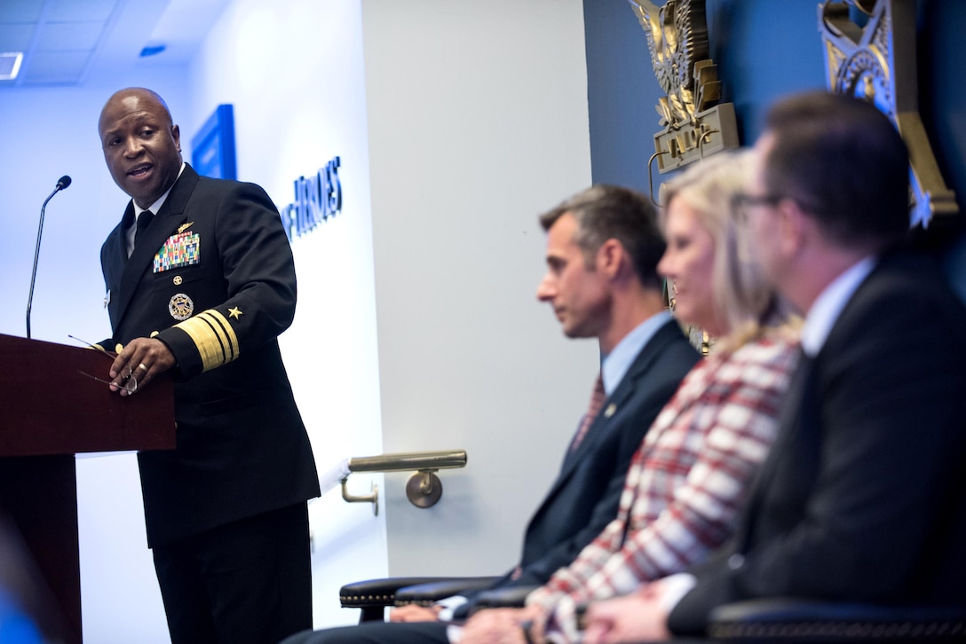 A person at a podium looks left at a row of people in chairs.