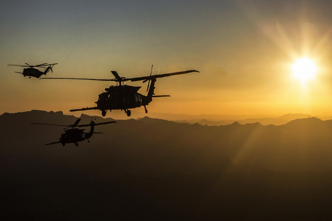 Three helicopters prepare for a refueling exercise with a low sun over the mountains.