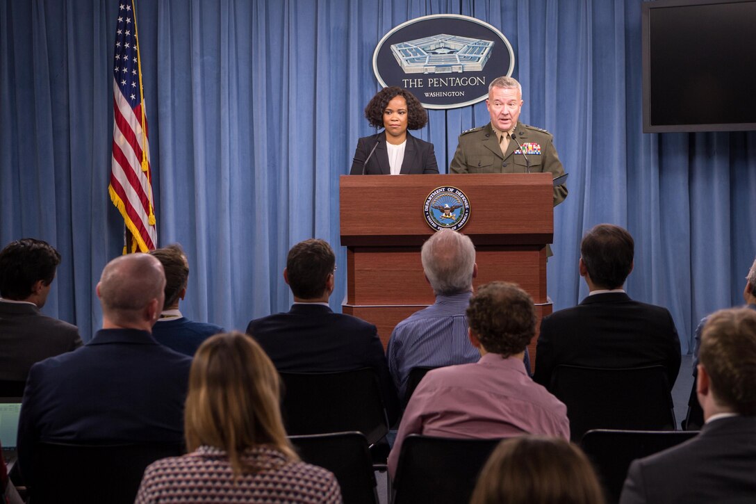 A civilian and a Marine general stand behind a podium speaking to a seated group.