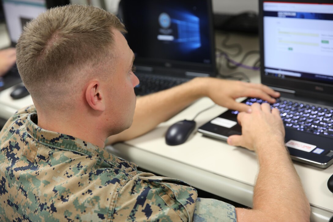 A soldier sits at a table and looks at a laptop screen with his hands on the keyboard.