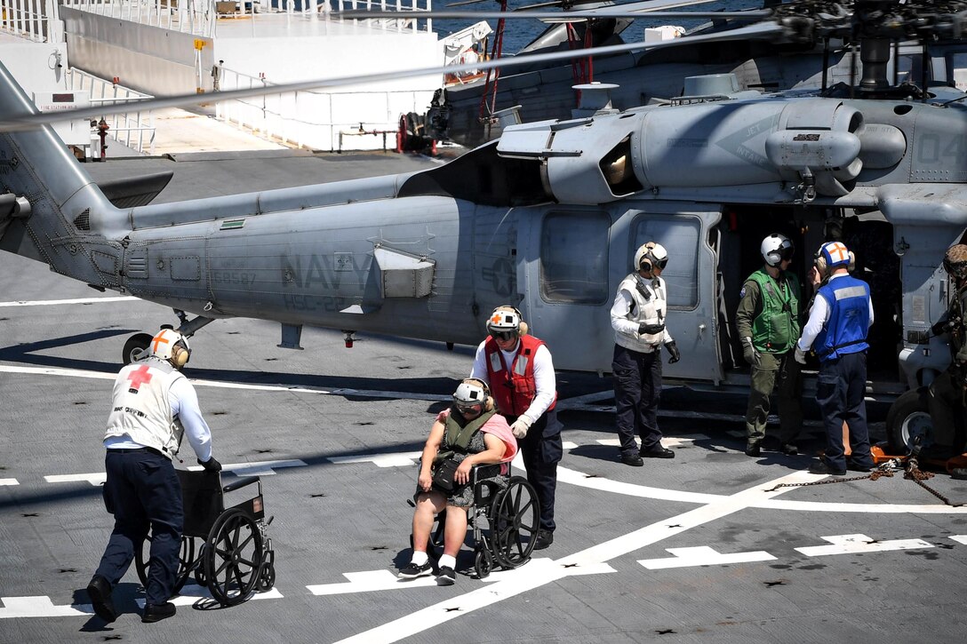 A sailor pushes a person in a wheelchair away from a helicopter.