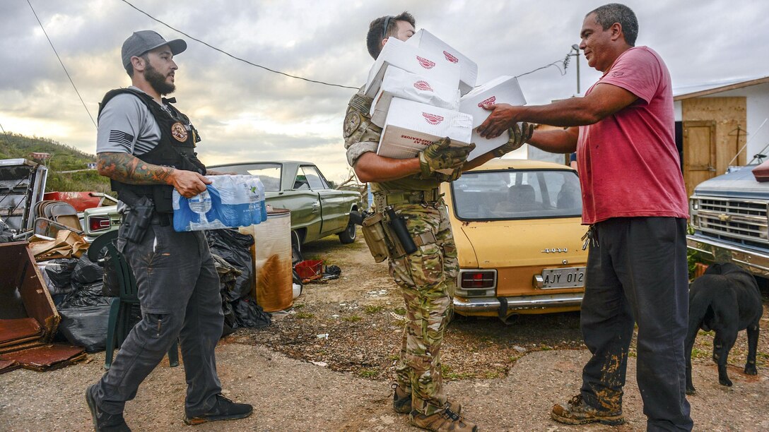 A man passes cardboard boxes to another man, while a third holds a case of water.