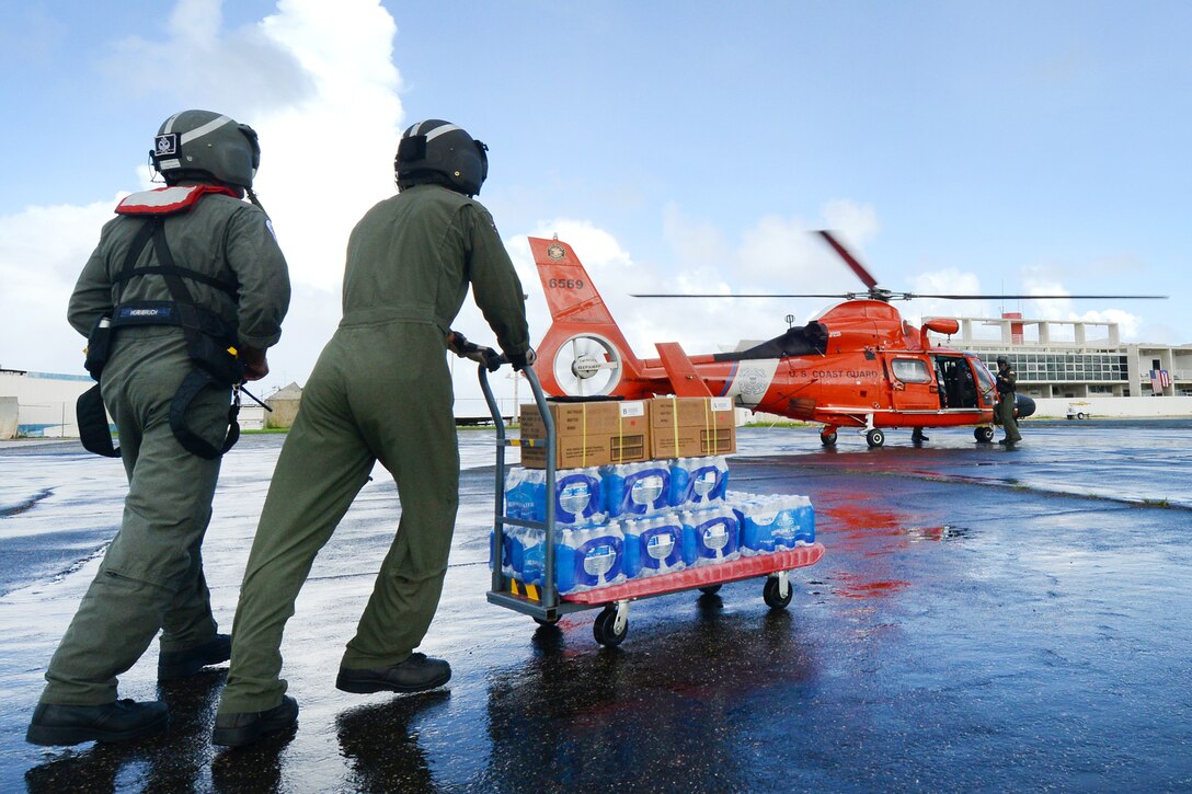 Two Coast Guardsmen push cases of water and boxes toward a helicopter.
