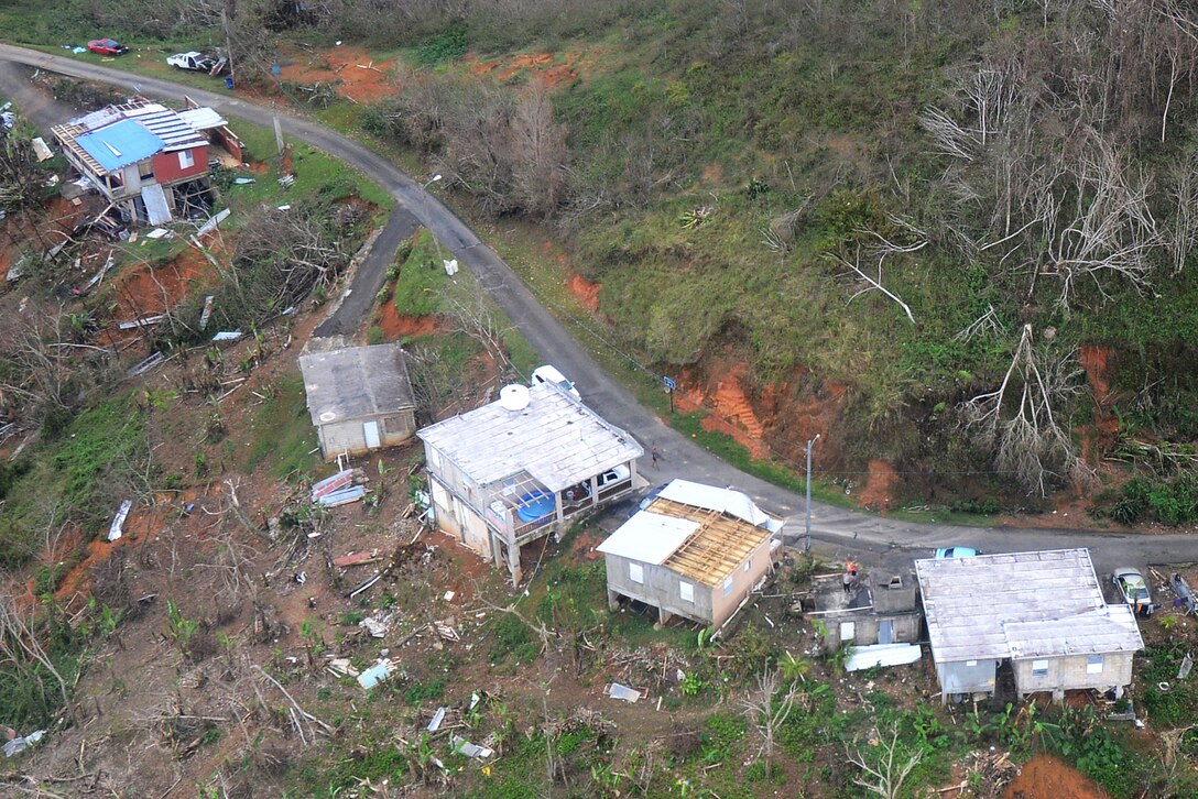 A row of damaged houses sit near a road with two people standing on the roof of one of them.