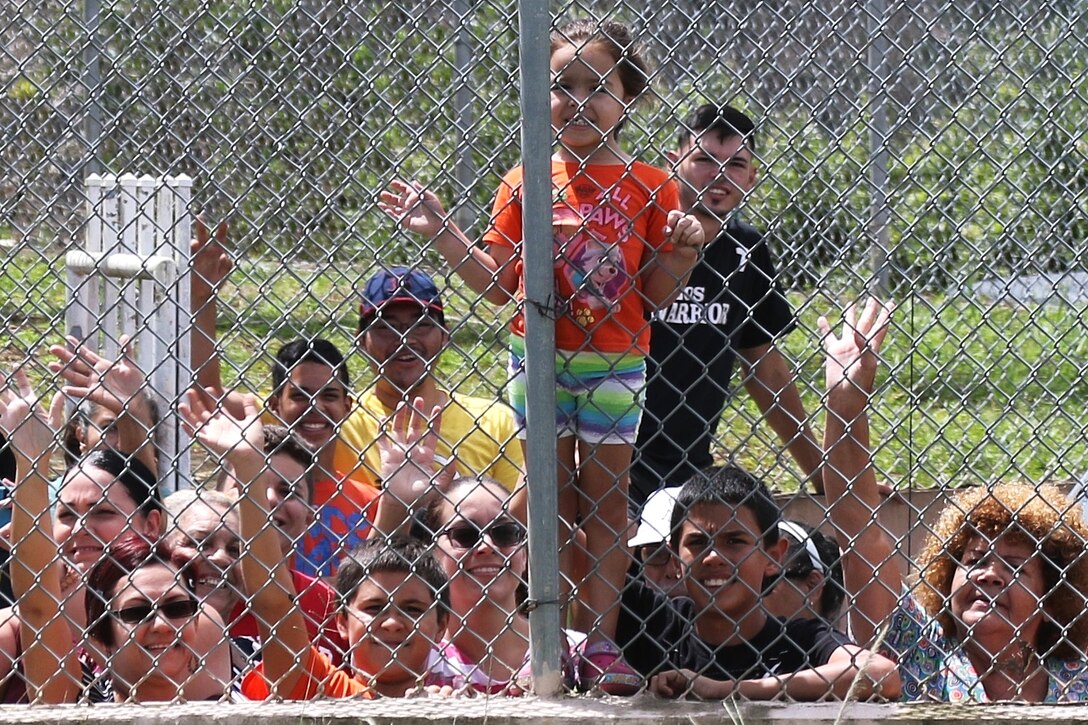 A child stands on a chain linked fence surrounded by other people.