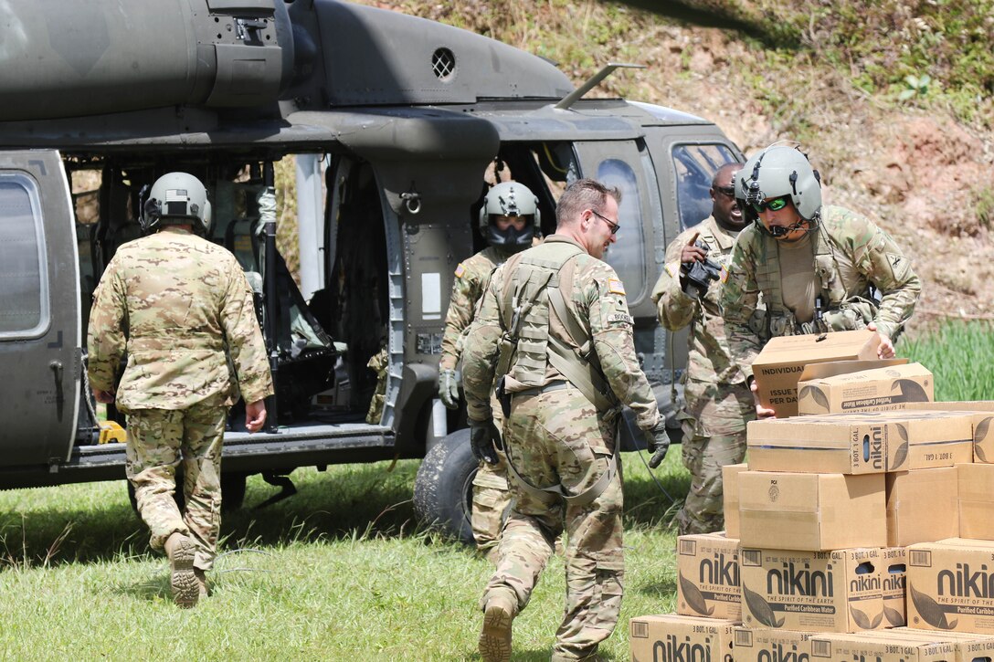 Soldiers remove boxes from a helicopter and put them into a pile on the ground.
