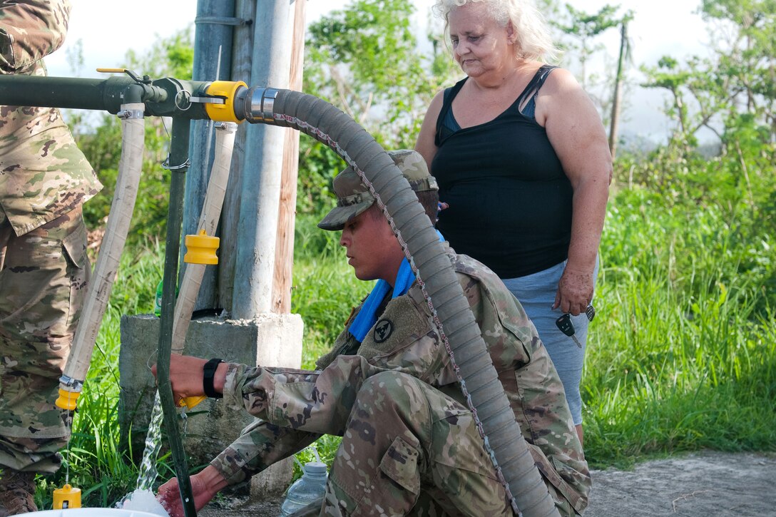 A soldier kneels while filling a bottle from a pipe with a civilian nearby.
